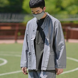 Puede incluir: Una persona vestida con una chaqueta gris con cuello mandarín, botones y bolsillos delanteros. La persona también lleva una mascarilla gris y una camiseta gris oscura. El atuendo se completa con pantalones grises a juego.