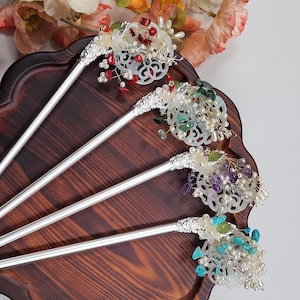 May include: A collection of silver hair sticks with ornate, decorative tops. Each stick features a unique arrangement of colorful beads, pearls, and gemstones in shades of red, green, purple, and turquoise. The hair accessories are displayed on a dark wooden tray.