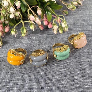 May include: Four gold-toned rings with colorful, stacked stone designs. The rings feature yellow, gray, green, and pink stones, each with a decorative gold band. The rings are displayed on a textured gray surface, with floral accents in the background.