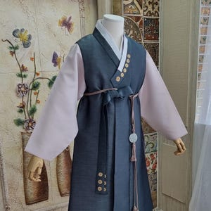 May include: A traditional Korean Hanbok dress in shades of teal, light pink, and white. The dress features a long skirt, a vest-like top with gold-colored button details, and long sleeves. A matching sash and decorative pendant complete the ensemble.