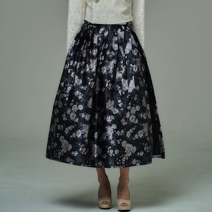 Puede incluir: Falda negra con estampado floral y diseño amplio y plisado. La falda presenta un patrón de flores rosas, azules y blancas. La modelo lleva tacones beige. La falda llega a la rodilla.