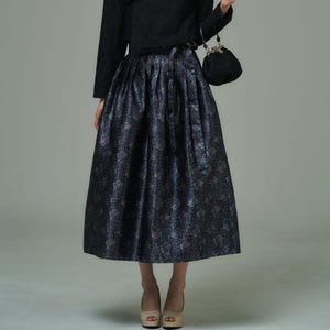 Puede incluir: Una falda midi azul marino oscuro con un estampado floral. La falda es plisada y tiene un ligero brillo. La modelo también lleva un top negro y un pequeño bolso negro.