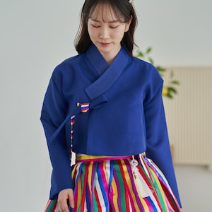 May include: A traditional Korean Hanbok outfit. The top is a solid blue with a V-neck and long sleeves. The skirt is a vibrant mix of vertical stripes in various colors, including red, yellow, green, and purple. A colorful ribbon detail accents the top.