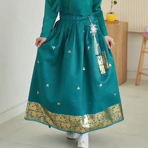 Puede incluir: Un vestido hanbok coreano tradicional de color turquesa. El vestido presenta una falda larga plisada con detalles florales dorados y un borde dorado. La parte superior es de color turquesa a juego con mangas largas. Un accesorio decorativo cuelga de la cintura.