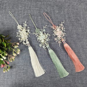 Op de afbeelding: Drie decoratieve kwasten met bloemaccenten. Elke kwast heeft een andere kleur: wit, groen en koraal. De kwasten hebben zilverkleurige metalen accenten en zijn gerangschikt op een grijze stoffen ondergrond.