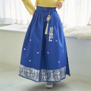 Puede incluir: Una falda azul vibrante con un dobladillo y detalles plateados con estampado floral. La falda presenta una cintura alta, pliegues y una corbata decorativa con borla. Pequeños detalles florales blancos están dispersos por la tela.
