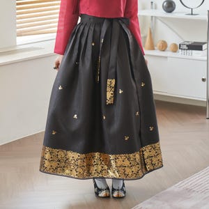 Puede incluir: Una falda negra con detalles florales dorados y una faja a juego. La falda tiene un borde floral dorado ancho en el dobladillo y pequeños detalles florales dorados. La parte superior es de color rojo liso.
