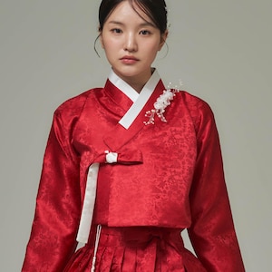 May include: A red traditional Korean Hanbok dress with a white collar and accents. The long-sleeved top features a floral pattern and a white bow. A white floral hair accessory is pinned to the top. The skirt is pleated and matches the top.