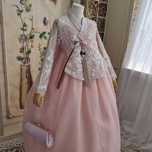 Puede incluir: Un vestido hanbok coreano tradicional en rosa suave. La parte superior presenta un intrincado encaje blanco sobre un corpiño rosa, mangas largas y una falda rosa a juego. Un pequeño bolso de mano lavanda es sostenido por el maniquí.