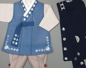 May include: A traditional Korean hanbok set featuring a blue vest with silver floral patterns, white long sleeves, and light beige trousers with red ties at the ankles. A matching dark navy headpiece with silver floral and character embroidery is included.