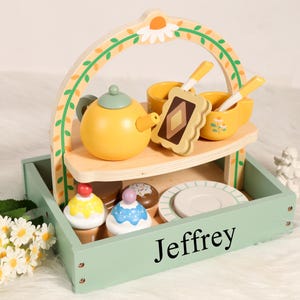 Könnte beinhalten: Ein hölzernes Spielzeug-Teeservice in einer hellgrünen Ablage mit Griff. Das Set enthält eine gelbe Teekanne, Tassen, Löffel, einen Teller und Cupcakes. Die Ablage ist mit "Jeffrey" beschriftet.