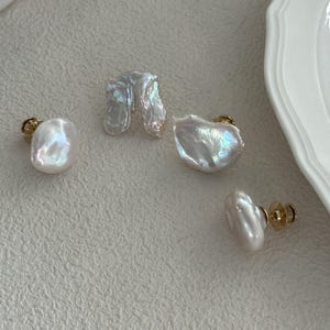 May include: Four white pearl earrings with gold posts are displayed on a textured surface. The pearls have irregular shapes and iridescent surfaces, reflecting light. The earrings vary in size and design, showcasing natural pearl formations.