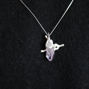 Puede incluir: Un collar de plata con un colgante que presenta un cristal de amatista en bruto. El colgante tiene un diseño abstracto único con un cristal de color púrpura claro. El collar se muestra sobre un fondo oscuro.