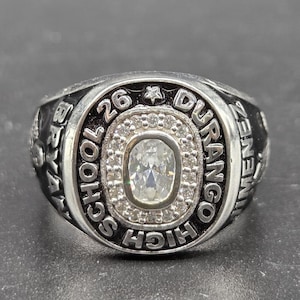 Könnte beinhalten: Silberner Abschlussring mit einem zentralen ovalen, klaren Stein, umgeben von kleineren Steinen. Der Ring trägt die Inschrift "DURANGO HIGH SCHOOL 26" und andere Texte. Ein Stern ist über der Zahl 26 zu sehen.
