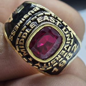 May include: A gold-colored ring with a large, square, ruby-red gemstone at the center. The ring has black enamel detailing with the words "Middle East University Business" and the year "96" inscribed.