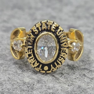 May include: A gold-colored ring with a central oval-shaped clear stone. The ring features the words "Tarleton State University" in black lettering around the central stone. Two smaller clear stones are set on either side of the ring.