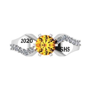 May include: A silver ring with a large, round, yellow gemstone at the center. The ring features a wave design with small, clear stones and the text "2020" and "SHS".