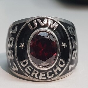 May include: Silver ring featuring a large, oval, deep red gemstone. The ring has a black band with the text "UVM DERECHO" and the numbers "87" and "88" in white. Two small star shapes are also visible.