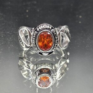 May include: Silver-toned class ring with an oval, orange-colored stone. The ring features the words "Senior High School" around the stone and the year "2026" on one side. The ring is reflected in the shiny surface below.