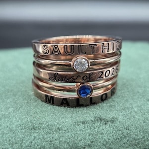 Puede incluir: Pila de anillos de oro rosa con texto grabado y piedras preciosas. Los anillos muestran las palabras "SAULT HIGH", "Class of 2026" y "MALLOR". Un anillo tiene una gema transparente y otro una gema azul.