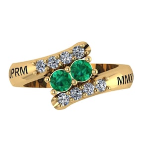 Könnte beinhalten: Ein goldfarbener Ring mit zwei smaragdgrünen Edelsteinen und sechs kleineren, klaren Edelsteinen. Der Ring trägt die Buchstaben "PRM" und "MMX" auf dem Band.