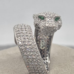 May include: A silver-toned wrap ring featuring a panther head design completely encrusted with small clear stones. The panther has two marquise-cut green stones for eyes. The band is wide and also covered in clear stones, creating a sparkling effect.