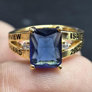 May include: A gold-colored ring featuring a large, rectangular, blue gemstone as the centerpiece. The ring has two smaller clear stones on either side of the main stone. The band is engraved with the words "IRVVIEW GUARD" and "ISABELLA 2023".