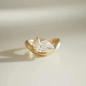May include: A gold ring featuring a pear-shaped diamond. The diamond is set in a bezel setting, and the band has a smooth, rounded design. The ring is displayed on a white surface.
