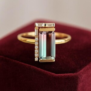 May include: A gold ring with a rectangular, multi-coloured gemstone in shades of pink, green, and brown. The ring has a vertical design with a small row of diamonds and a rectangular top. The ring is set on a red velvet surface.