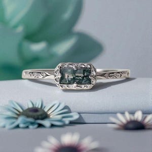 May include: A silver ring featuring a rectangular moss agate gemstone. The stone is set in an ornate bezel. The band has an engraved floral design. The ring is displayed on a light blue surface with blue flowers in the foreground.