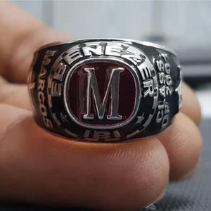 May include: A silver and black class ring with a large red centre featuring a capital "M". The ring is inscribed with "MARCOS EBENEZER CLASS 2007" around the edges. The ring is held by a hand.