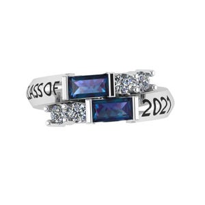 May include: A silver ring with the text "CLASS OF 2021". The ring features two rectangular, dark blue gemstones and several smaller, clear gemstones. The ring has a modern, elegant design.
