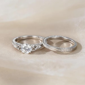 Puede incluir: Dos anillos de plata. El anillo de compromiso presenta una piedra preciosa grande, redonda y transparente y detalles intrincados de nudos celtas. La alianza a juego tiene un delicado patrón grabado. Ambos anillos están sobre un fondo suave y neutro.