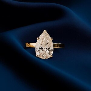 May include: A gold ring with a large, pear-shaped diamond. The diamond is set with four prongs and sparkles against a dark blue background. The band is a simple, polished gold.