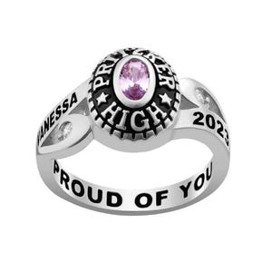 May include: A silver class ring with a central oval pink stone. The ring is inscribed with "Vanessa", "Proud of You", "Premier High", and "2023" in black. Small clear stones are set on the ring's shoulders.