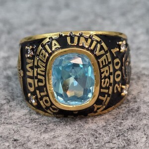 May include: A gold-toned ring with a large, light blue gemstone at the center. The ring has a black band with the words "COLUMBIA UNIVERSITY" and "MD 2002" inscribed. Small, clear stones are set on the sides.