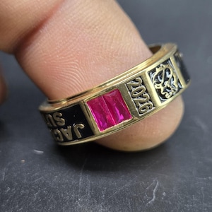 Puede incluir: Un anillo dorado con una piedra rectangular de color magenta. El anillo tiene secciones de esmalte negro con la inscripción "JAC SU" y el año "2026". Hay patrones decorativos grabados en la superficie del anillo.