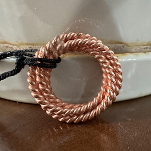 Puede incluir: Un colgante circular de color cobre con un diseño de cuerda trenzada. El colgante está sujeto a un cordón negro. El colgante se muestra sobre un fondo borroso.