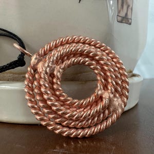 May include: A copper-colored pendant necklace featuring a circular design made of twisted metal. The pendant is suspended from a black cord. The necklace is displayed against a neutral background.