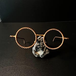 May include: A pair of round, copper-colored wire-framed glasses with a twisted rope design around the lenses. The glasses are resting on a dark gray rock against a black background. The glasses have a vintage or steampunk aesthetic.