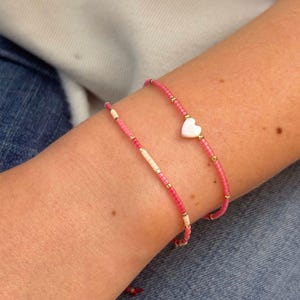 Könnte beinhalten: Zwei zarte Perlenarmbänder aus kleinen rosafarbenen Saatperlen, die mit winzigen goldfarbenen Metallzwischenperlen durchsetzt sind. Ein Armband hat ein einzelnes weißes herzförmiges Element als Mittelpunkt, während das andere einen Abschnitt mit cremefarbenen Perlen aufweist.