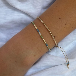 May include: Two delicate beaded bracelets. One bracelet features a single strand of small, uniform gold-toned metallic beads. The second bracelet consists of a mix of tiny beads in shades of blue, green, cream, and gold, with two thin dangling cords.
