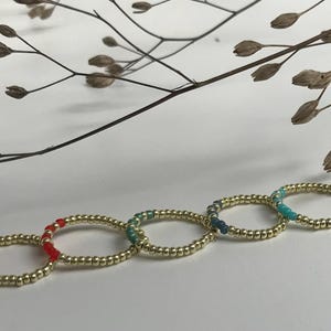 Könnte beinhalten: Fünf goldfarbene Ringe mit farbigen Perlen in Rosa, Rot, Türkis, Blau und Türkis. Die Ringe liegen auf einer weißen Oberfläche, im Hintergrund ein Zweig mit getrockneten Pflanzen.