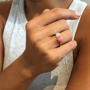 Op de afbeelding: Een gouden ring met een lichtroze en wit edelsteenontwerp. De ring heeft een gedraaide band en twee ronde edelstenen, één lichtroze en één wit. De ring wordt aan een vinger gedragen.