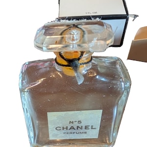 Puede incluir: Un frasco de perfume Chanel No. 5 vintage con un diseño claro y geométrico. El frasco tiene un tapón transparente y una etiqueta con el nombre de la marca y el nombre del perfume. La caja es blanca con texto negro. El frasco es de 1 fl oz.