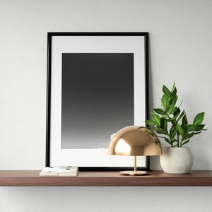 Puede incluir: Una impresión en blanco y negro enmarcada, una lámpara de escritorio dorada y una planta en maceta con hojas verdes se encuentran en una estantería marrón. La lámpara tiene una pantalla redondeada y una base delgada. La planta está en una maceta blanca.