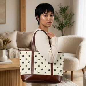 以下が含まれることがあります： クリーム色のトートバッグ。黒い花柄が繰り返されています。茶色の革のハンドルと茶色の革のベースが付いています。バッグは人が持っています。