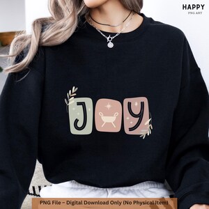 May include: Black sweatshirt featuring the word "JOY" in a decorative font. The "J" is in a green square, the "O" has a manger graphic in a pink square, and the "Y" is in a pink square. The text "HAPPY PNG ART" is in the upper right corner.