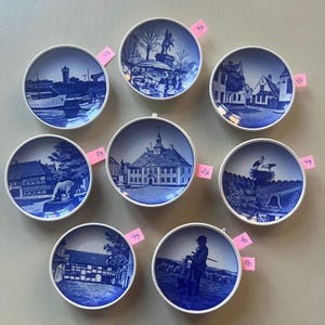 Può includere: Una collezione di otto piatti decorativi blu e bianchi. Ogni piatto presenta un'illustrazione scenica diversa, tra cui edifici, paesaggi e figure. I piatti hanno un bordo bianco e sono probabilmente in porcellana o ceramica. Alcuni piatti hanno etichette rosa con i prezzi.