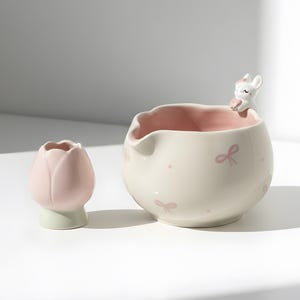 Może przedstawiać: Dwa ceramiczne elementy dekoracyjne. Jeden to wazon w kształcie tulipana w kolorze różowym i zielonym. Drugi to kremowa miska z różowym wnętrzem, ozdobiona różowymi kokardkami i małą figurką zwierzątka na brzegu.